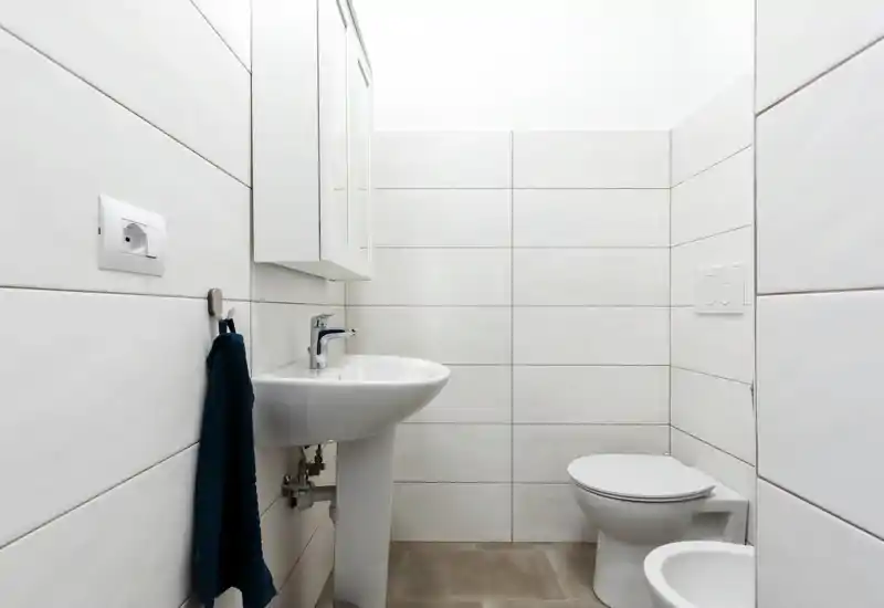 Compact, modern white bathroom with wall-mounted sink, toilet and bidet, tiled walls and a navy hand towel—clean and minimal.
