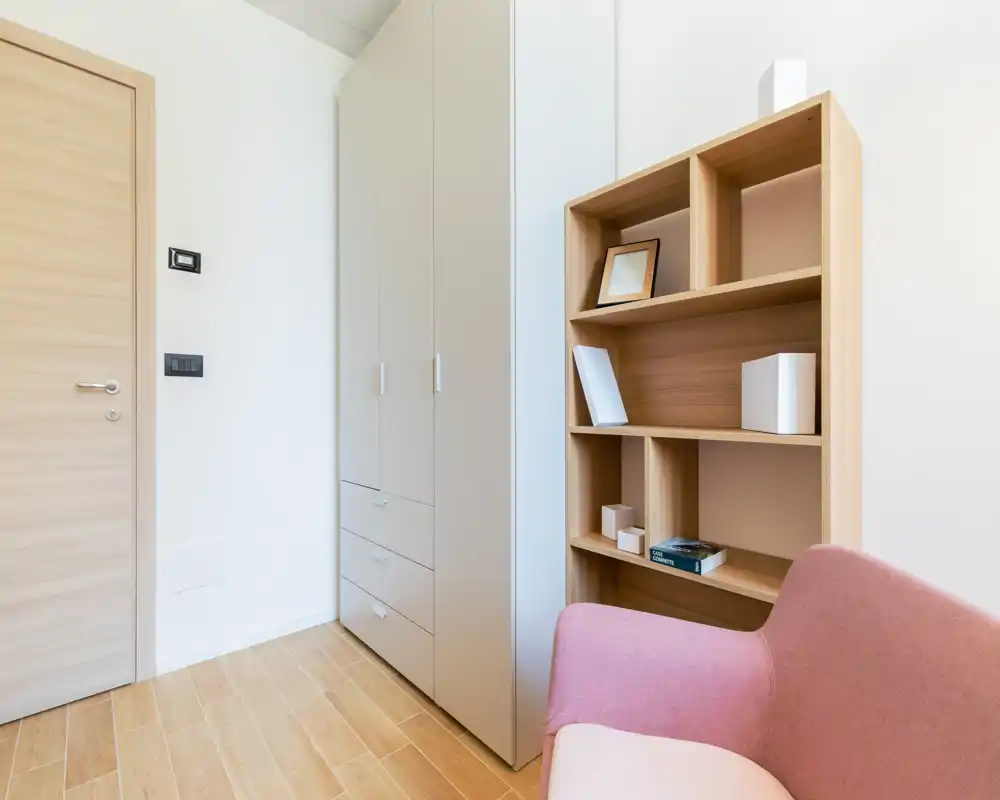 Bright, minimal living area corner with a pink armchair, wooden shelving and a tall wardrobe — neat, contemporary styling suitable for rental listings.