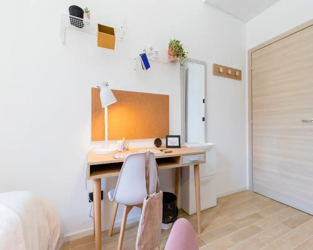 Bright, minimalist bedroom corner with a wooden desk, cork noticeboard and a small portion of the bed visible at the edge. The space features warm wood tones, a desk lamp and plants for a cozy study nook.
