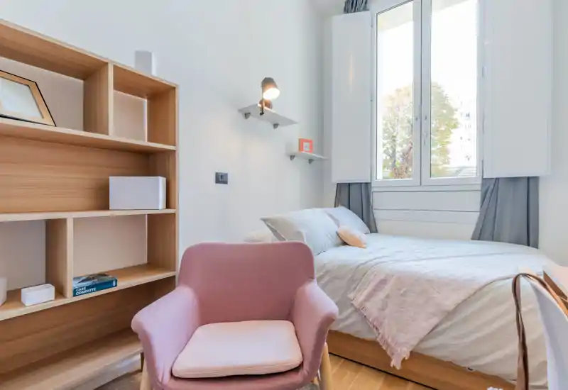 Bright, modern bedroom with a large bed dressed in soft linens, a cozy pink armchair in the foreground and built-in shelving. Natural light enters through a tall window, creating a welcoming, well-styled space ideal for rentals.