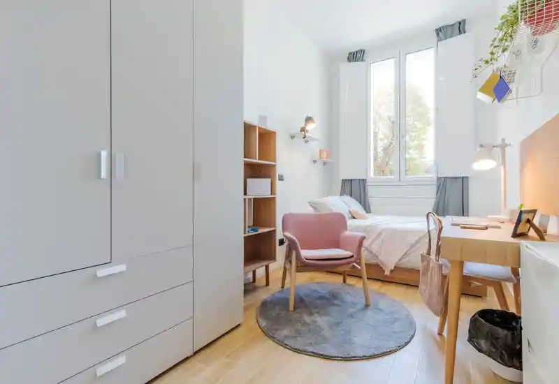 Bright, stylish single bedroom with a comfortable bed beneath a large window, a cozy pink armchair on a round rug, built-in wardrobe and a wooden desk for studying or remote work.