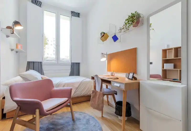 Sunlit, well-staged single bedroom with a comfortable bed, wooden desk and cozy armchair — ideal for studying or relaxing.