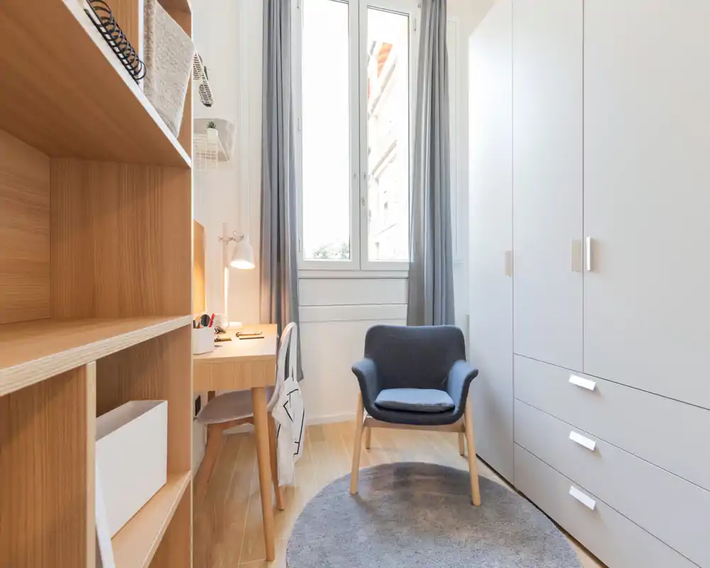 Rincón de estar moderno y luminoso con sillón tapizado, alfombra redonda y un gran armario. La luz natural de la ventana alta y las estanterías de madera cálida crean un rincón acogedor, perfecto para relajarse o leer.