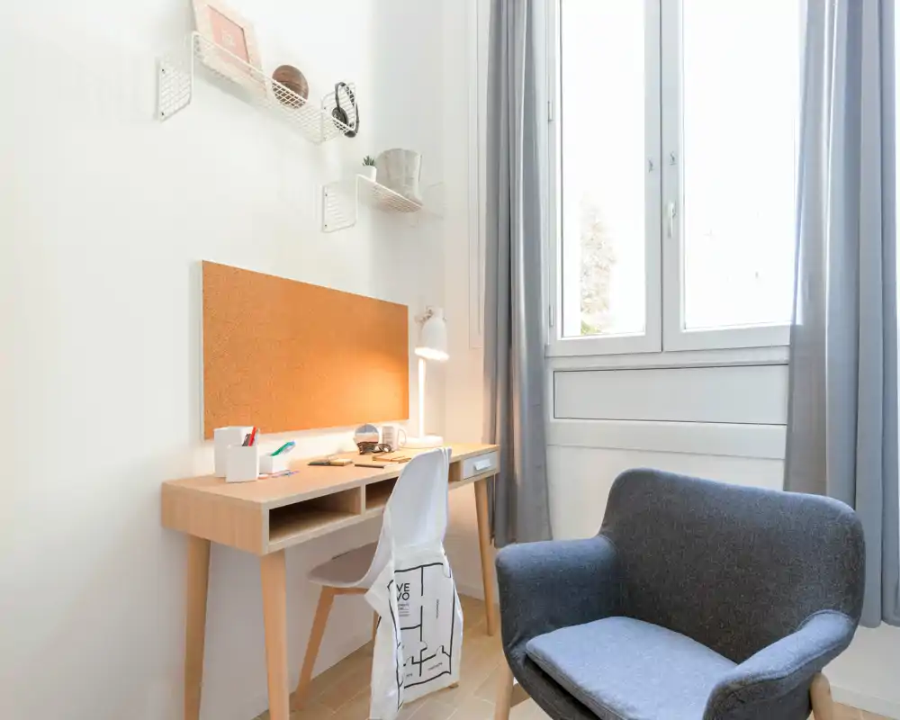 Rincón de dormitorio luminoso y minimalista con escritorio de madera, tablero de corcho y un sillón acogedor junto a una gran ventana — ideal para estudiar o leer.