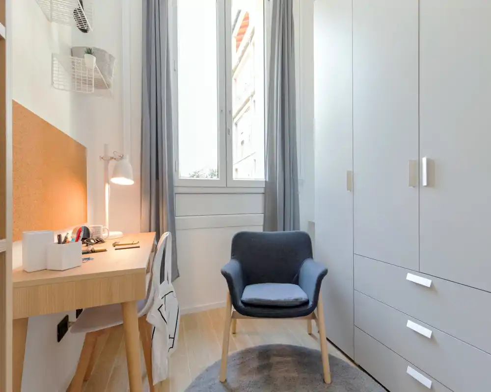 Rincón de dormitorio pequeño y luminoso con escritorio, un sillón cómodo y un gran armario junto a una ventana alta que aporta luz natural.