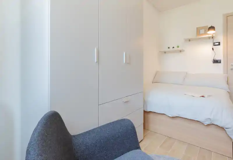 Dormitorio compacto y luminoso con una cama individual bien hecha, base de madera clara y ropa de cama en tonos neutros. Un armario blanco alto y un sillón acogedor completan el ambiente moderno y sereno.