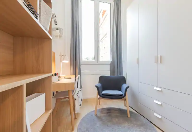 Small furnished room with a wardrobe, shelving, a desk and a comfortable armchair by a window. Neutral tones, wooden flooring and soft natural light create a calm study or dressing area.