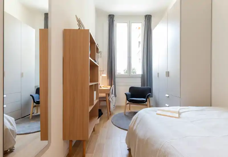 Dormitorio luminoso y bien decorado con cama arreglada, estantería de madera, butaca acogedora y amplio armario; la luz natural de la ventana realza la decoración moderna y limpia.