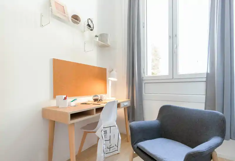 Rincón de dormitorio luminoso y minimalista con escritorio de madera, tablero de corcho y un sillón acogedor junto a una gran ventana — ideal para estudiar o leer.