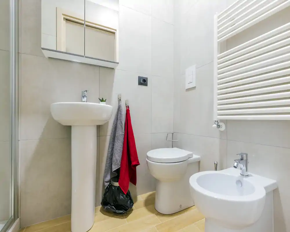 Baño moderno y compacto con lavabo de pedestal, inodoro y bidé blancos sobre paredes azulejadas claras y suelo efecto madera.