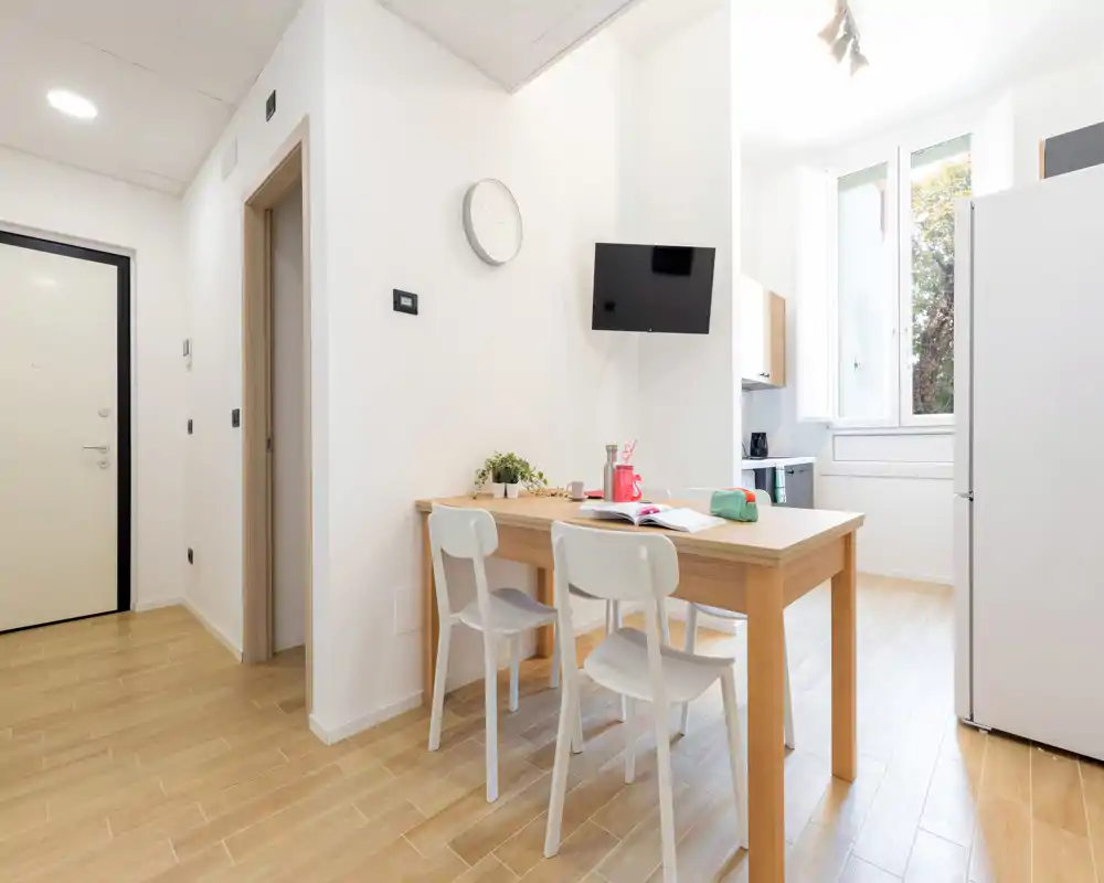 Zona de comedor moderna y luminosa con mesa de madera y dos sillas blancas junto a una gran ventana; decoración minimalista y luz natural perfectas para un apartamento compacto.