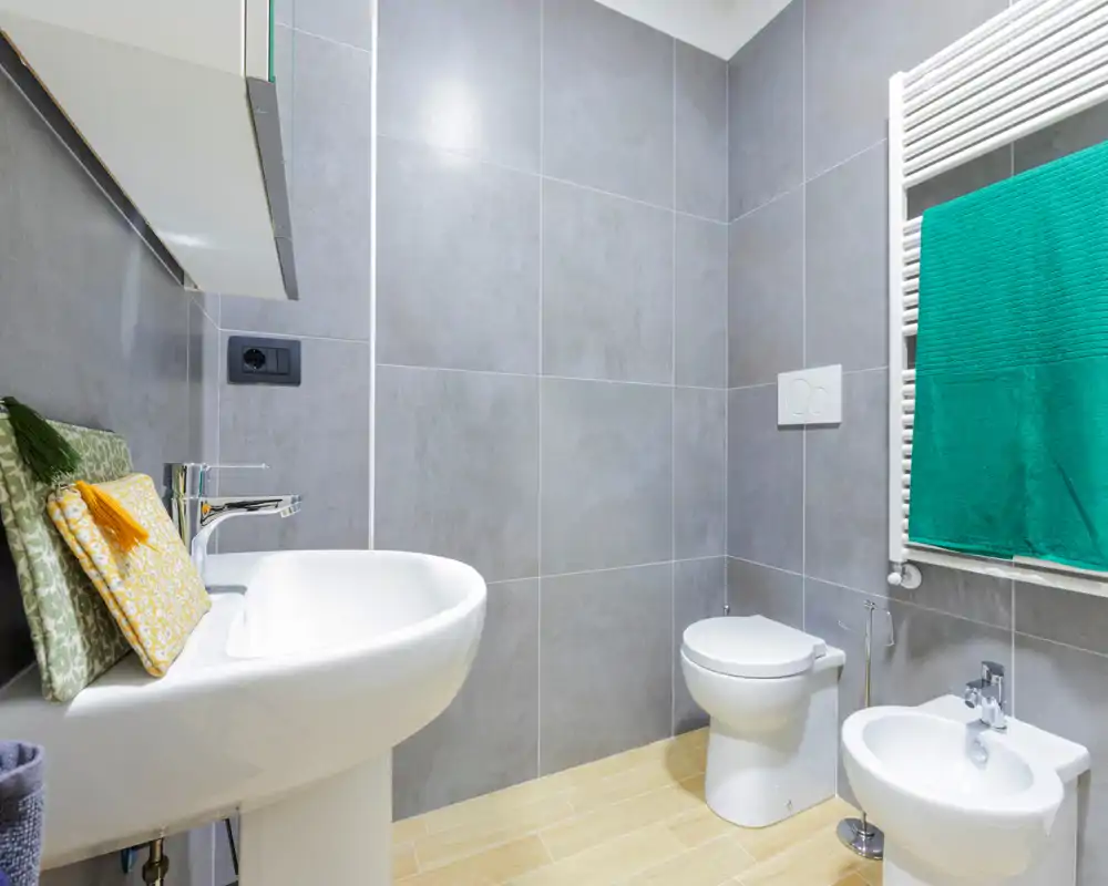 Baño moderno y bien iluminado con azulejos grises, lavabo de pie, inodoro y bidé, y toallero calefactado con toalla verde.