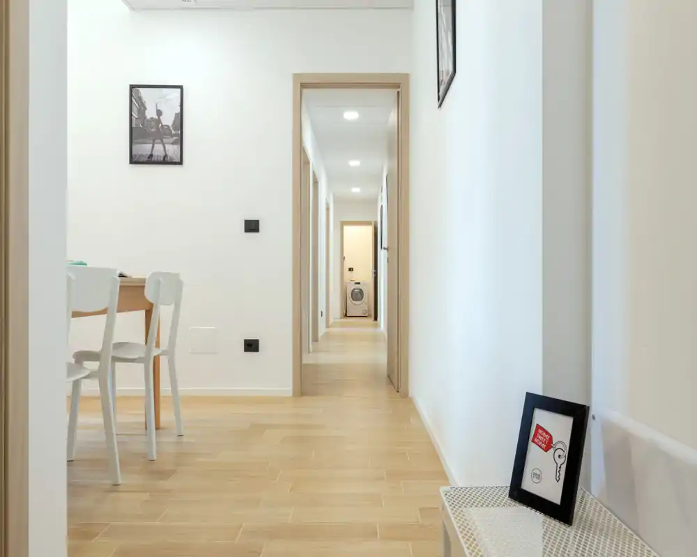 Entrada/pasillo luminoso y minimalista con suelo de madera clara y paredes blancas; pequeña consola con marco y vista hacia un pasillo bien iluminado que conduce al área de servicio.