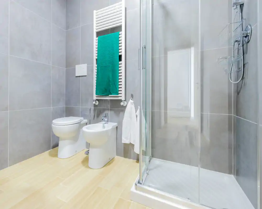 Baño moderno con mampara de ducha de cristal, radiador-toallero mural con toalla verde, bidé y WC sobre suelo efecto madera claro y grandes azulejos grises en las paredes.