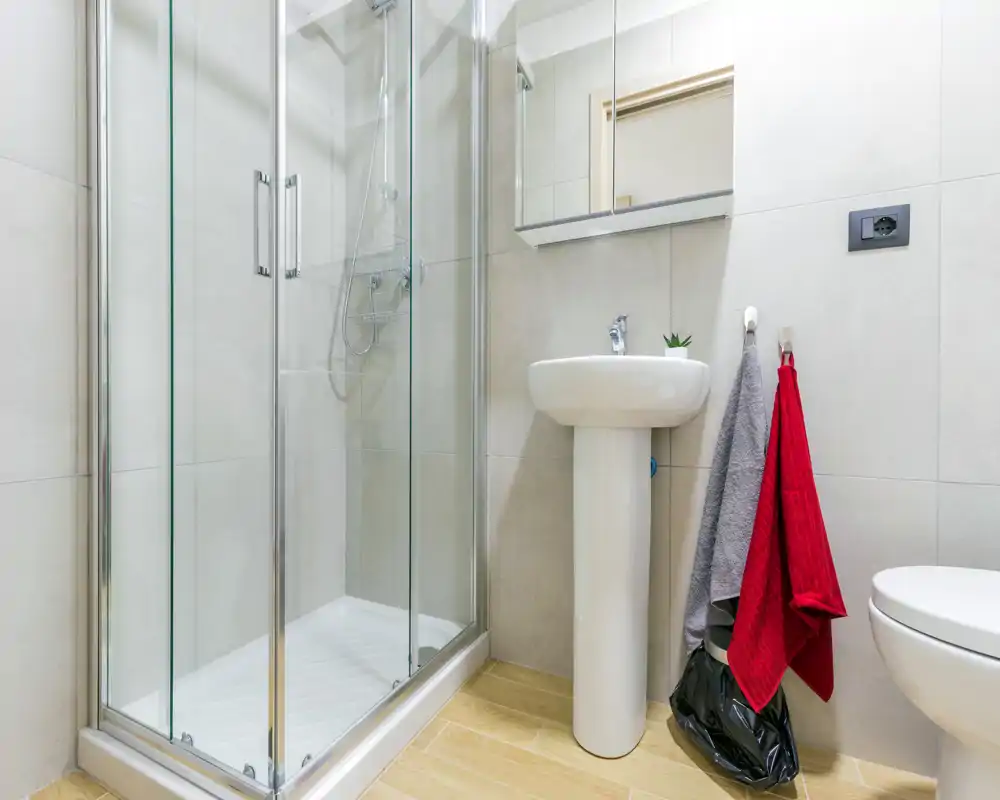 Baño moderno y luminoso con mampara de ducha de vidrio, lavabo exento, espejo y váter. Azulejos neutros y suelo efecto madera para una estética limpia y contemporánea.