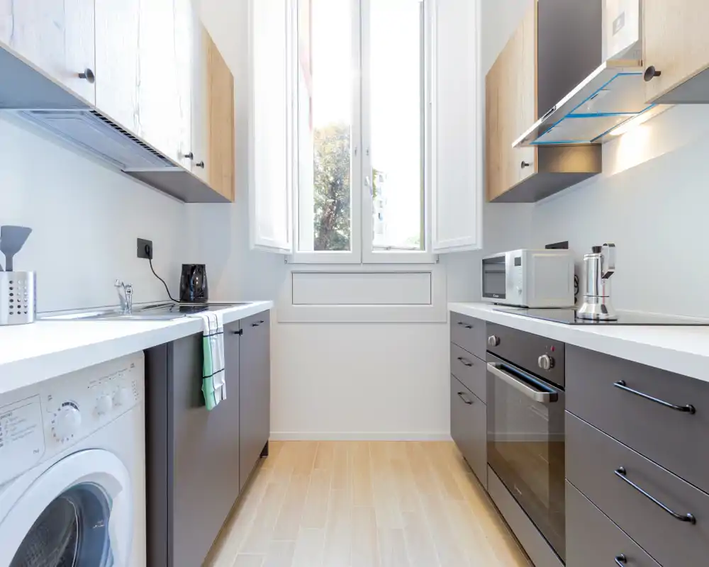 Cocina moderna en pasillo, luminosa, con electrodomésticos integrados, amplia ventana central y suelo efecto madera limpio. Perfecta para apartamentos urbanos compactos que buscan un espacio funcional y con estilo.
