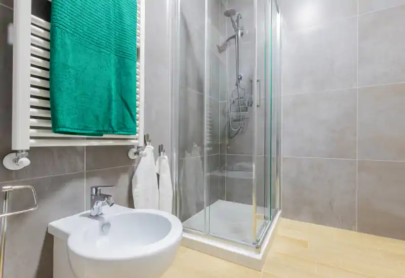 Modern bathroom with a glass shower enclosure, wall-mounted radiator with a green towel, sink and neutral tile finishes — bright and clean.
