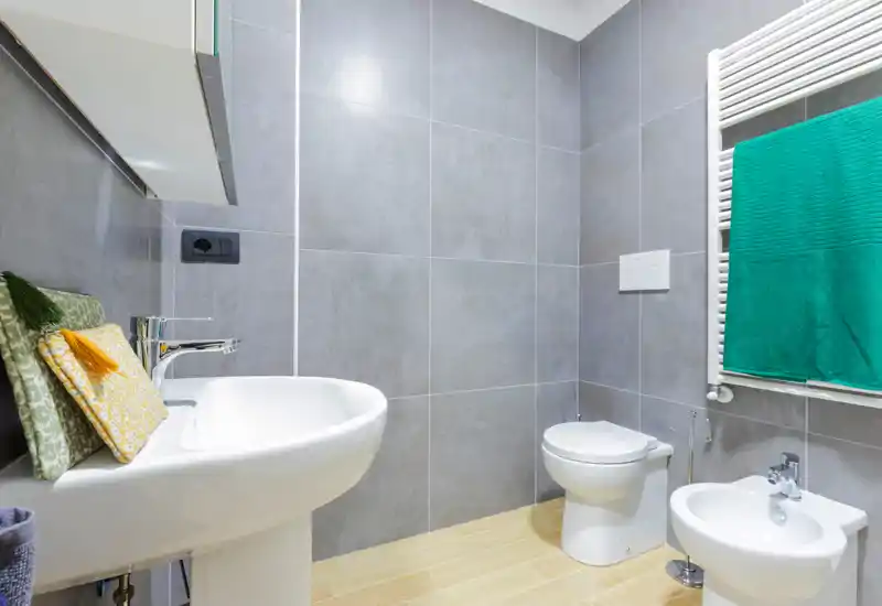 Modern bathroom with wall-mounted towel radiator, bidet and toilet, white sink and neutral grey tiles creating a clean, contemporary look.