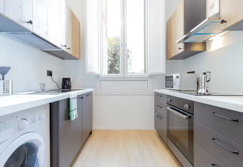 Bright modern kitchen with grey lower cabinets, wood-effect upper cabinets and a central window providing natural light. Includes built-in oven, microwave, countertop appliances and a washing machine—clean, functional space ideal for rental listings.