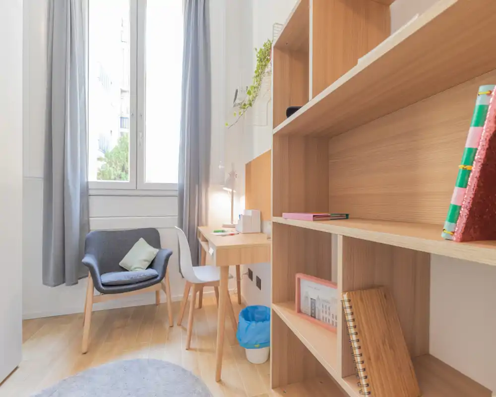 Coin chambre/étude lumineux et soigné avec bureau, fauteuil et étagères ; grande fenêtre apportant une belle lumière naturelle et une décoration neutre et apaisante.