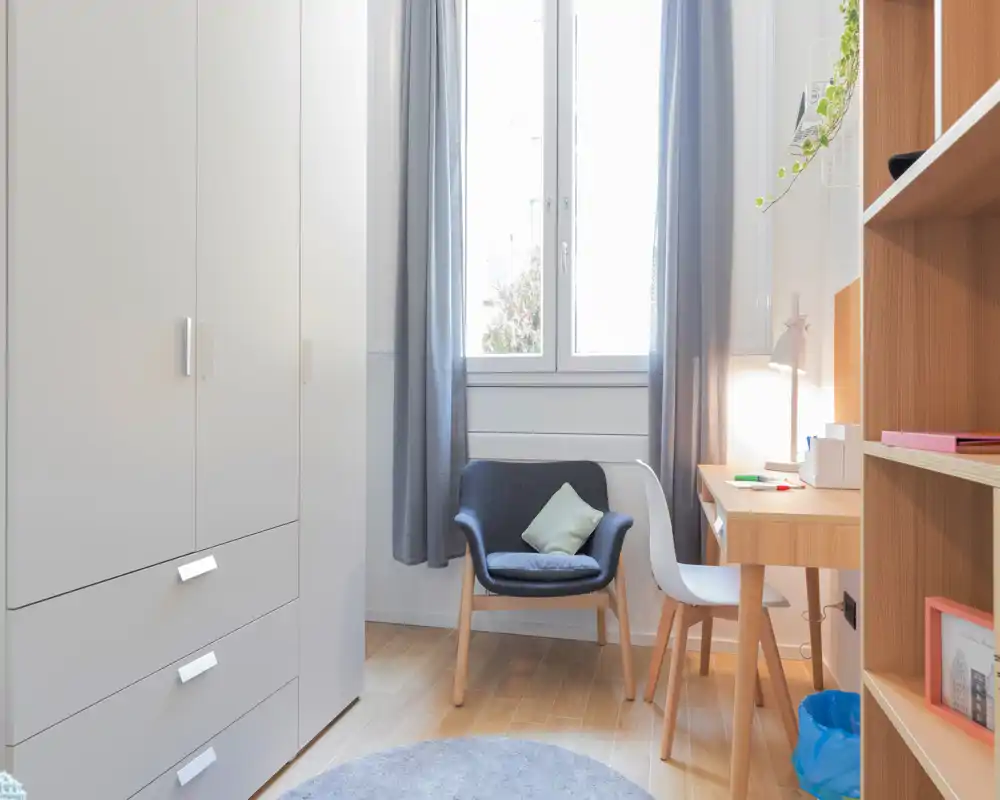 Coin de chambre petit et lumineux avec armoire, bureau et un fauteuil confortable près de la fenêtre. La lumière naturelle et les textiles doux apportent une ambiance apaisante.