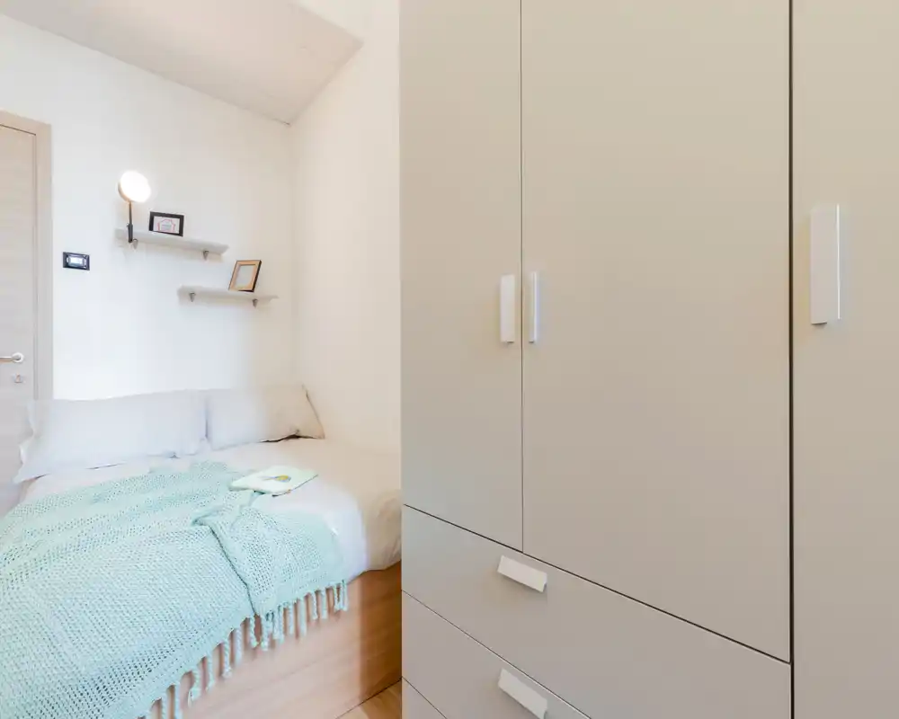 Chambre compacte et lumineuse avec un lit bien fait, coussins doux et un jeté vert menthe; une armoire intégrée occupe le premier plan pour le rangement.