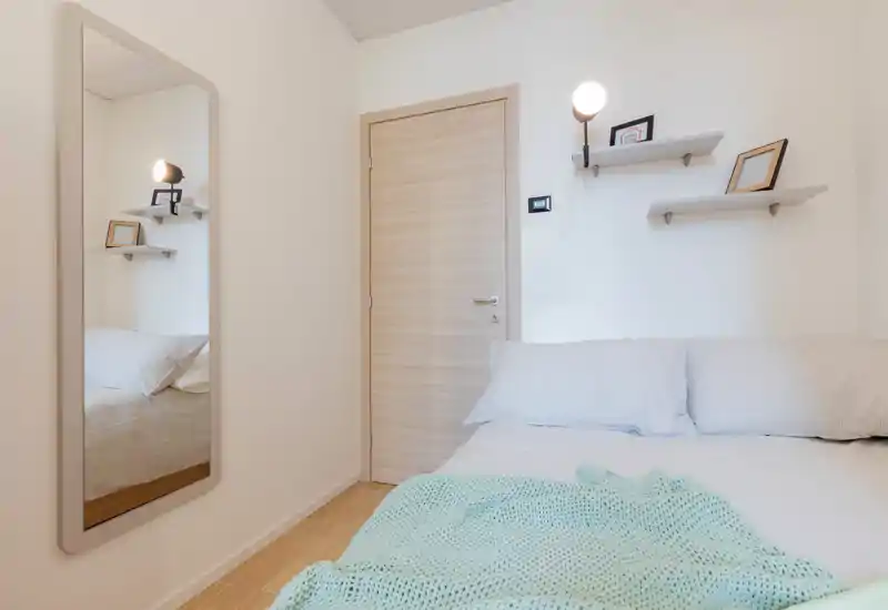Chambre lumineuse et minimaliste avec un lit double partiellement visible, une porte en bois clair, des étagères murales et une grande glace qui apporte de la profondeur.