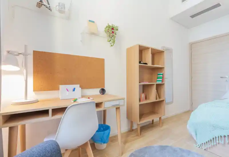 Coin chambre lumineux et moderne avec bureau en bois, étagère et lit partiellement visible avec couvre-lit clair—idéal pour un étudiant ou une personne seule.