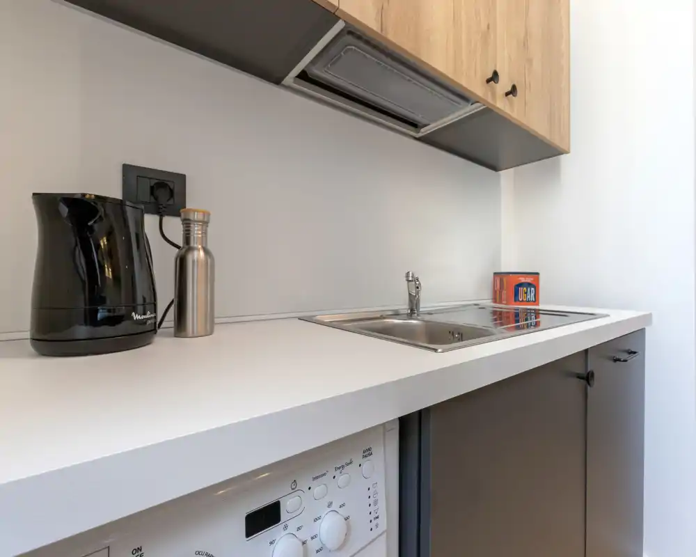 Kitchenette moderne et compacte avec évier inox, plan de travail blanc, bouilloire et hotte; agencement propre, lumineux et fonctionnel, adapté aux courts séjours.