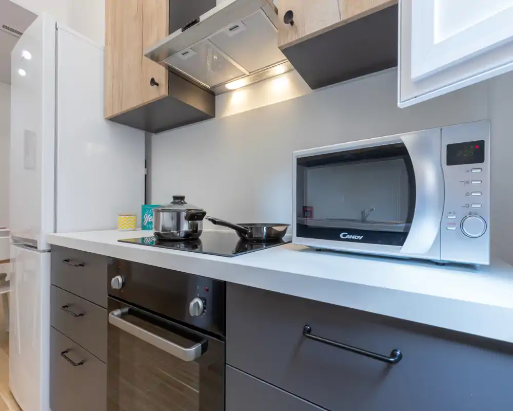 Cuisine moderne compacte avec four encastré, plaque à induction et micro-ondes sur un plan de travail blanc propre ; bien éclairée et soigneusement rangée pour la location.