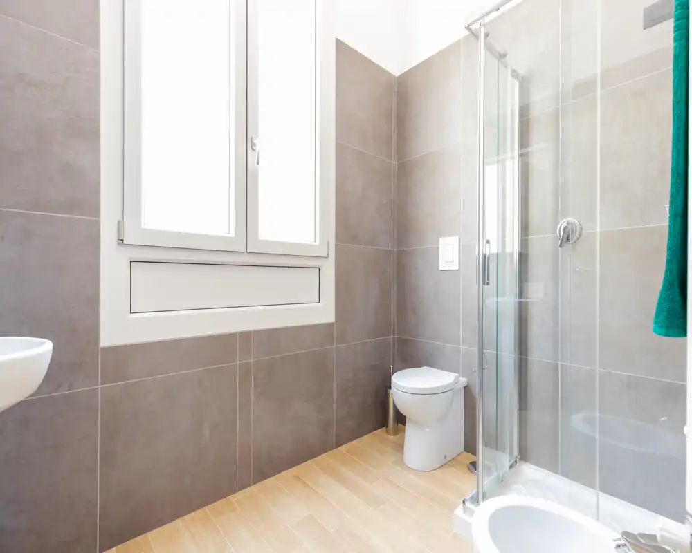 Salle de bains moderne et lumineuse avec douche vitrée, toilette murale et une grande fenêtre dépoli apportant la lumière naturelle.