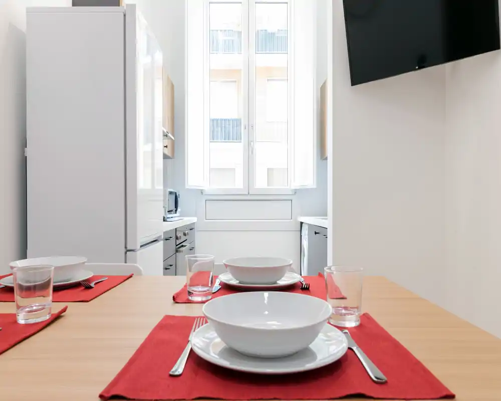 Cuisine moderne avec coin repas, table dressée pour quatre avec vaisselle blanche sur sets rouges et lumière naturelle venant d'une grande fenêtre; électroménagers compacts et style minimaliste.