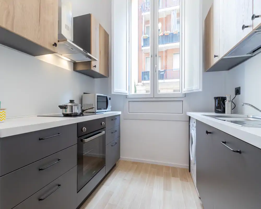 Cuisine moderne et lumineuse avec four encastré, plaque de cuisson, micro-ondes et une grande fenêtre apportant beaucoup de lumière; meubles épurés et sol effet bois pour un espace propre et accueillant.