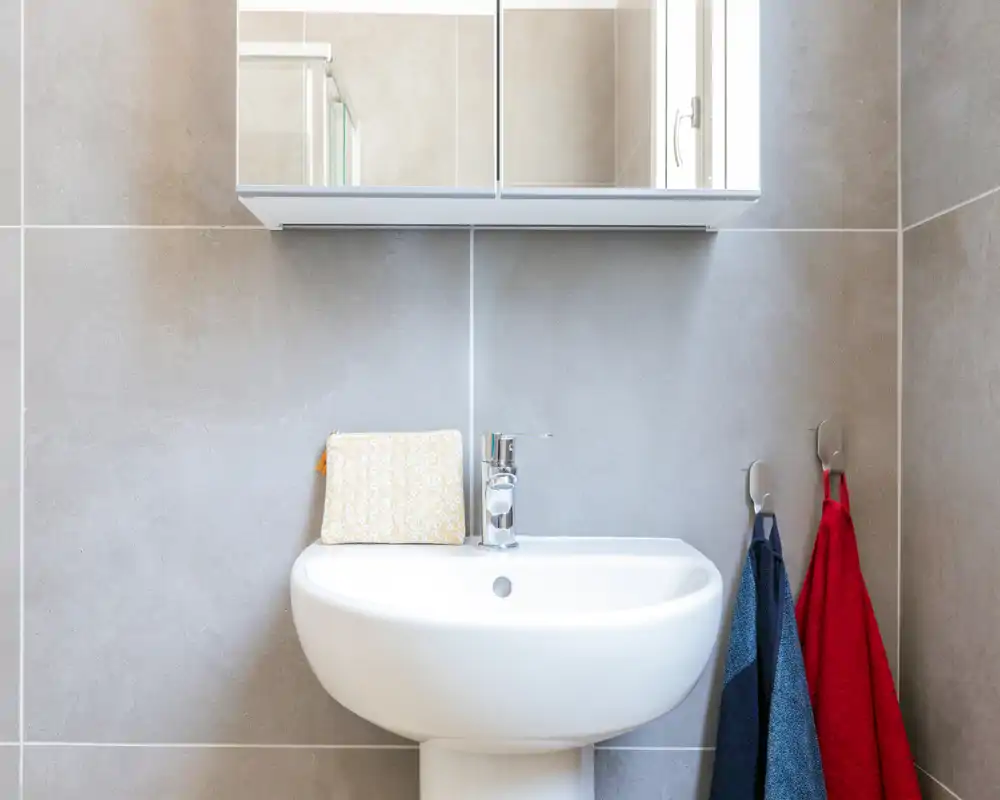 Salle de bain moderne et lumineuse avec lavabo sur pied blanc, armoire-miroir murale et carrelage gris neutre ; des serviettes colorées apportent une touche chaleureuse.