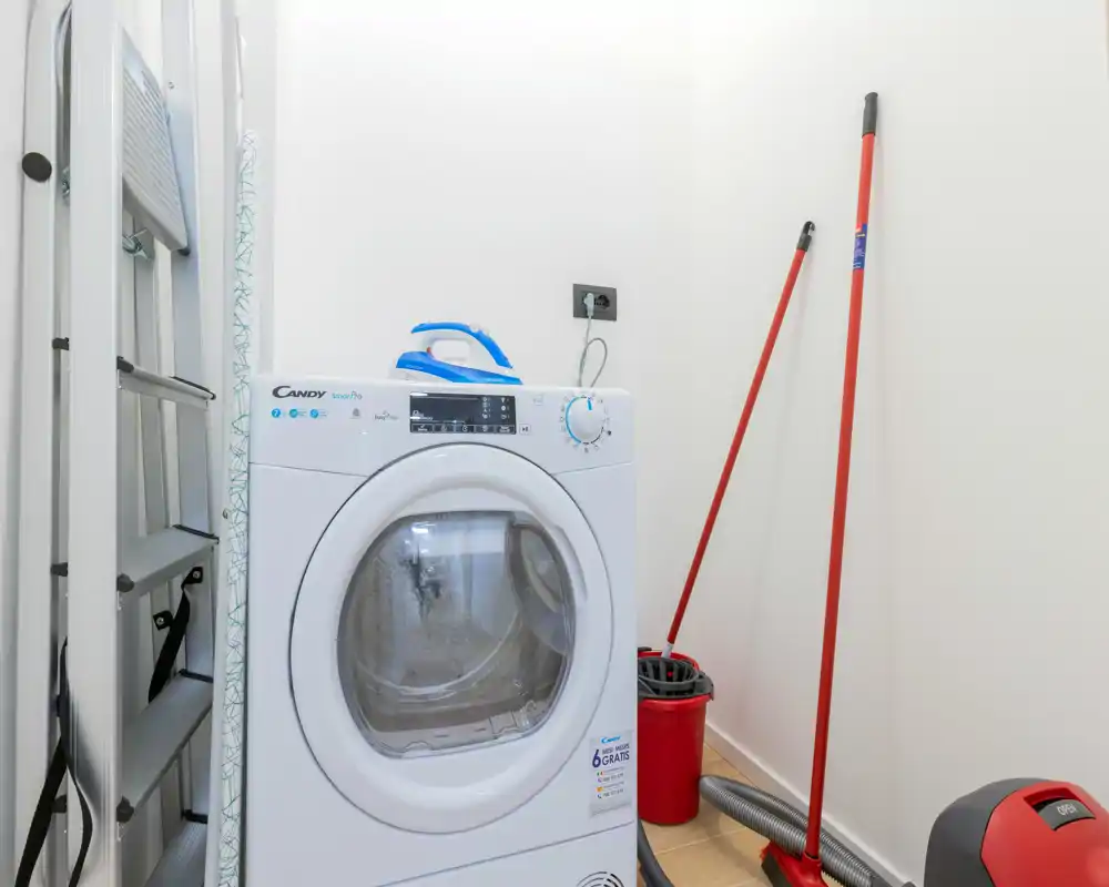 Petit coin buanderie/lingerie avec machine à laver à hublot, planche à repasser et outils de nettoyage. Murs blancs et agencement compact pour un rangement pratique.
