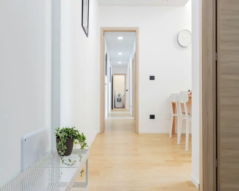 Entrée/couloir lumineux et minimaliste avec sol en bois clair, murs blancs et un petit banc avec une plante; une salle à manger est visible à droite et un long couloir mène aux autres pièces.