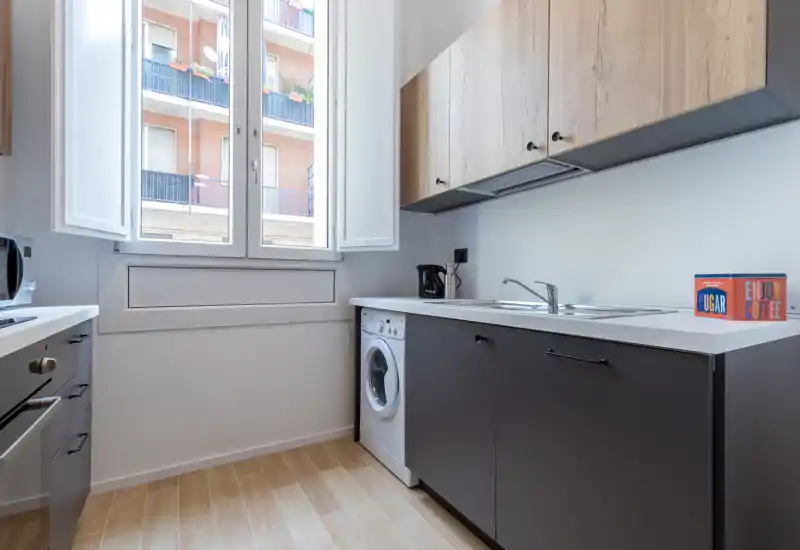 Bright modern kitchen with wood-accented upper cabinets, dark lower cabinetry, integrated sink and a washing machine under the counter. Large window provides abundant natural light and a clean, functional layout ideal for apartment listings.
