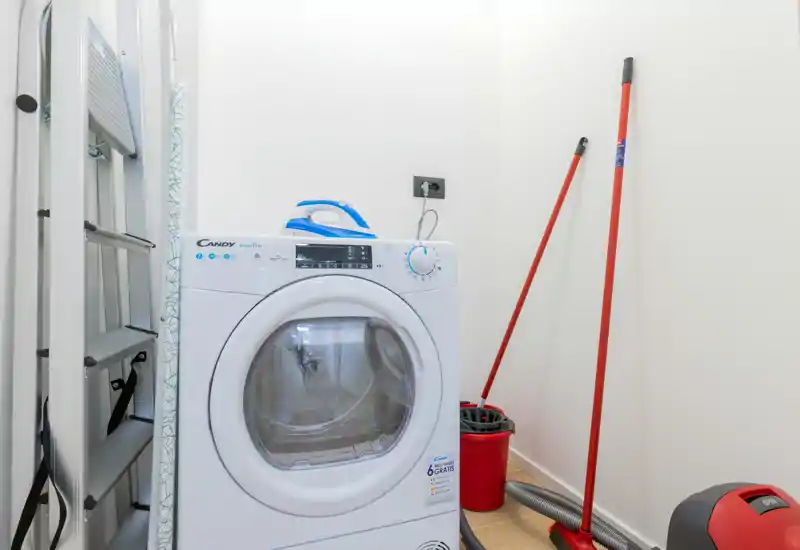 Small utility/laundry corner with a front-loading washing machine, ironing board and cleaning tools. Bright white walls and compact layout ideal for practical storage.