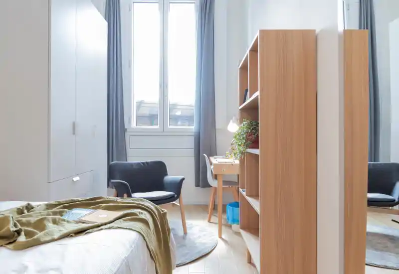 Dormitorio moderno y luminoso con cama visible, butaca y pequeño escritorio junto a una gran ventana; estantería de madera clara que aporta calidez.