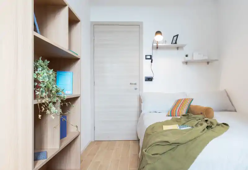 Dormitorio luminoso y minimalista con la cama bien hecha a la derecha, textiles suaves y una estantería de madera a la izquierda; estilo nórdico limpio, perfecto para un anuncio de alquiler.
