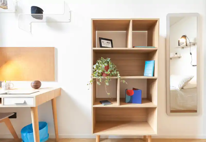Rincón de dormitorio luminoso y minimalista con una estantería de madera, escritorio y un espejo que refleja la cama. El espacio es amplio y bien iluminado, con detalles cálidos como una planta y una lámpara suave.