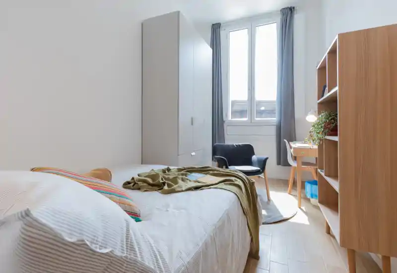 Dormitorio luminoso y bien decorado con una cama individual cómoda, luz natural procedente de una gran ventana, armario, escritorio y textiles acogedores.