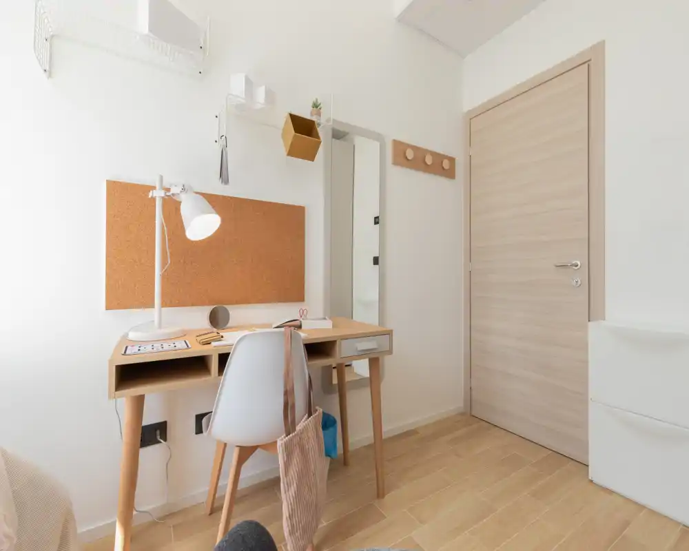 Coin chambre lumineux et minimaliste avec bureau en bois clair, panneau en liège et chaise blanche — espace de travail soigné adapté aux locations courtes.