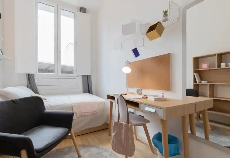 Chambre lumineuse et moderne avec un lit double, un bureau en bois et un fauteuil confortable ; textiles doux et fenêtre apportant lumière naturelle, idéale pour une annonce locative.