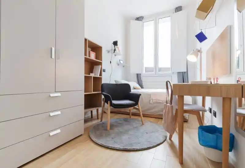 Chambre simple lumineuse et moderne avec lit devant une grande fenêtre, un fauteuil confortable sur un tapis rond et un bureau en bois parfait pour étudier ou travailler.