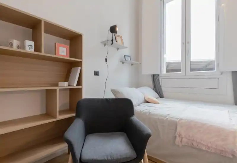 Chambre compacte et lumineuse avec un lit simple bien fait près d'une grande fenêtre, un fauteuil confortable et des étagères intégrées — idéale pour un séjour cosy en solo.