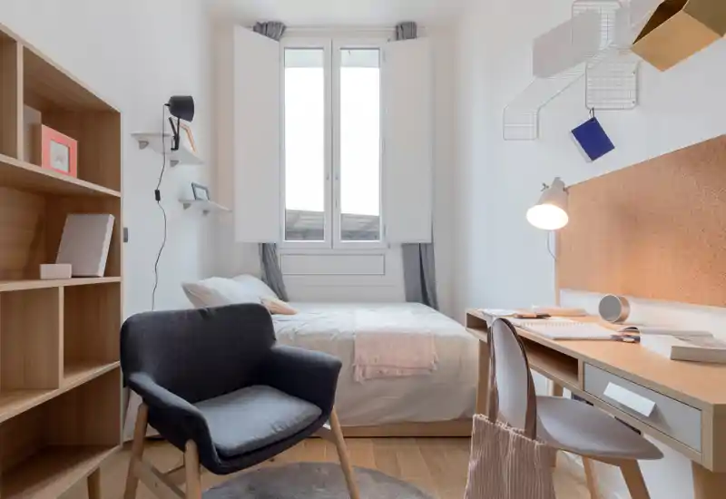 Chambre simple lumineuse et chaleureuse avec lit bien fait sous une grande fenêtre, bureau avec lampe et étagères organisées — idéale pour un étudiant ou une personne seule.