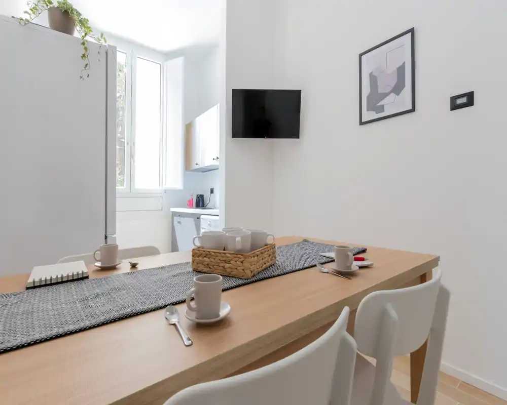 Coin repas lumineux et minimaliste avec une table en bois dressée pour le café, chaises blanches modernes et lumière naturelle provenant d’une grande fenêtre créant une ambiance chaleureuse.