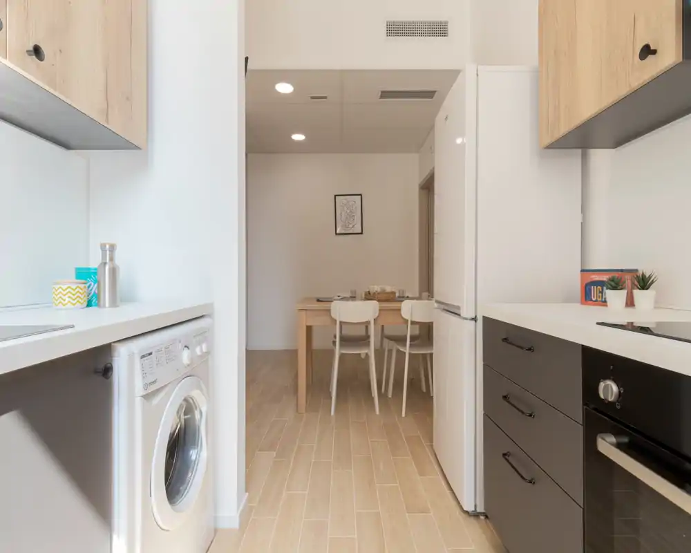 Cuisine compacte et moderne avec électroménagers intégrés, sol en bois clair et petit coin repas en arrière‑plan ; lumineuse, propre et bien décorée pour une annonce.