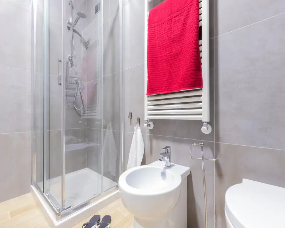 Salle de bains moderne et lumineuse avec cabine de douche en verre, bidet, WC et sèche-serviettes avec une serviette rouge. Carrelage gris neutre et sol effet bois pour une ambiance propre et contemporaine.